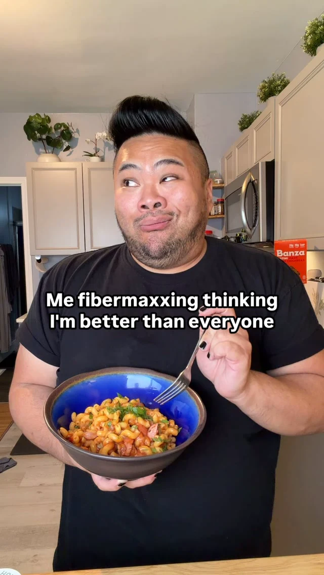 I hold a bowl of Banza pasta beneath text reading, Me fibermaxxing thinking I’m better than everyone, while a Banza box sits on the counter behind me.
