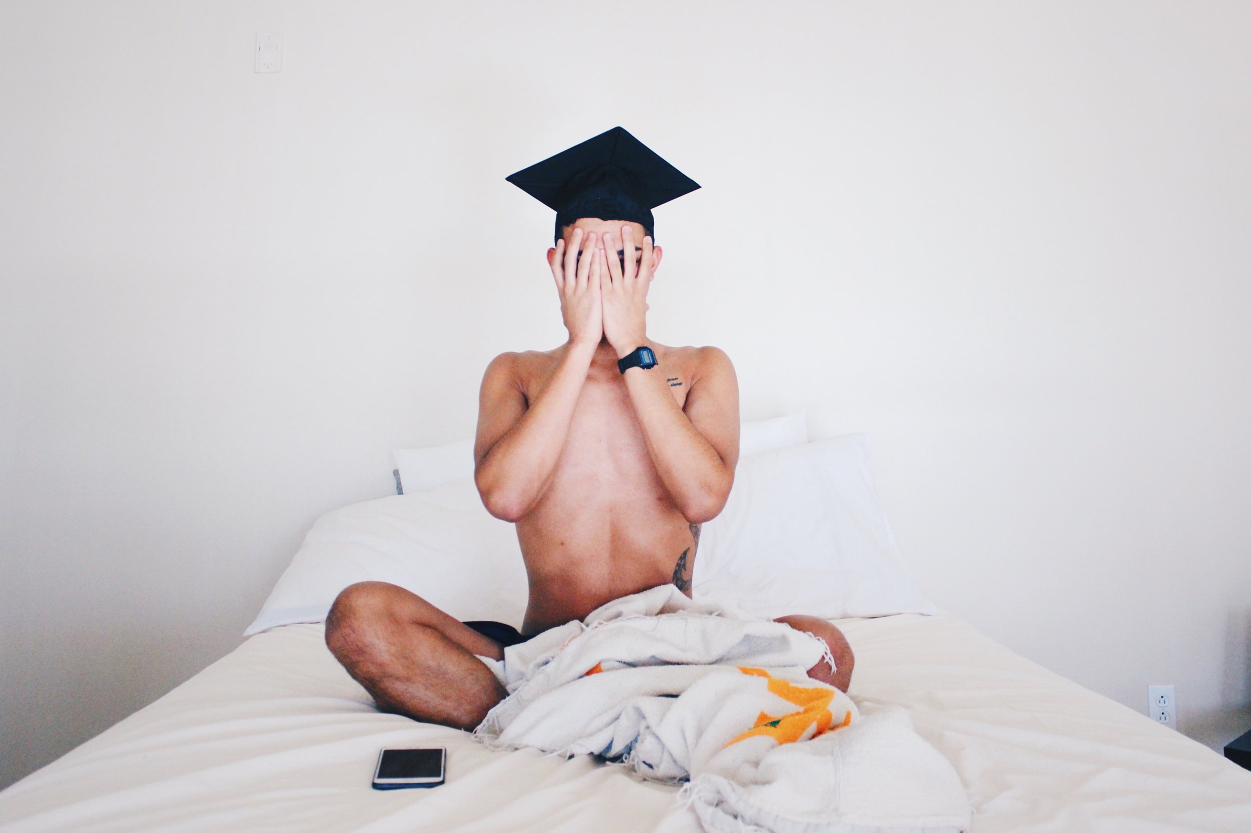 A graduate in a cap sitting on a bed with their face in their hands