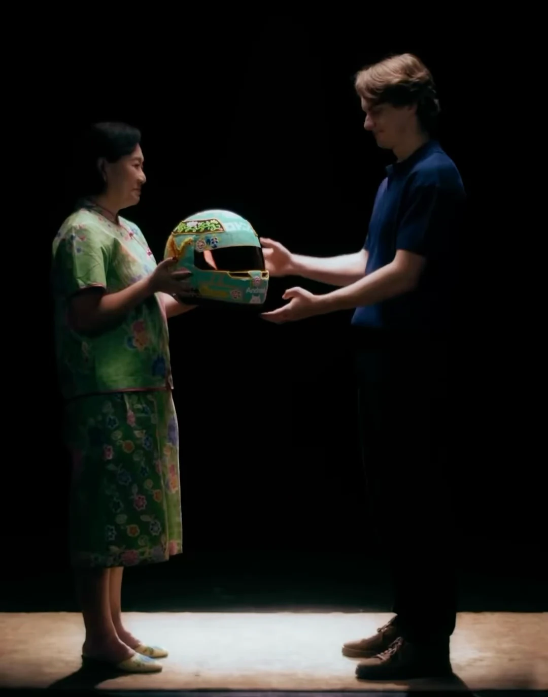 Angeline Kong and Oscar Piastri hold the turquoise beaded helmet between them against a black background.