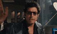 Ian Malcolm raises one hand while wearing sunglasses.