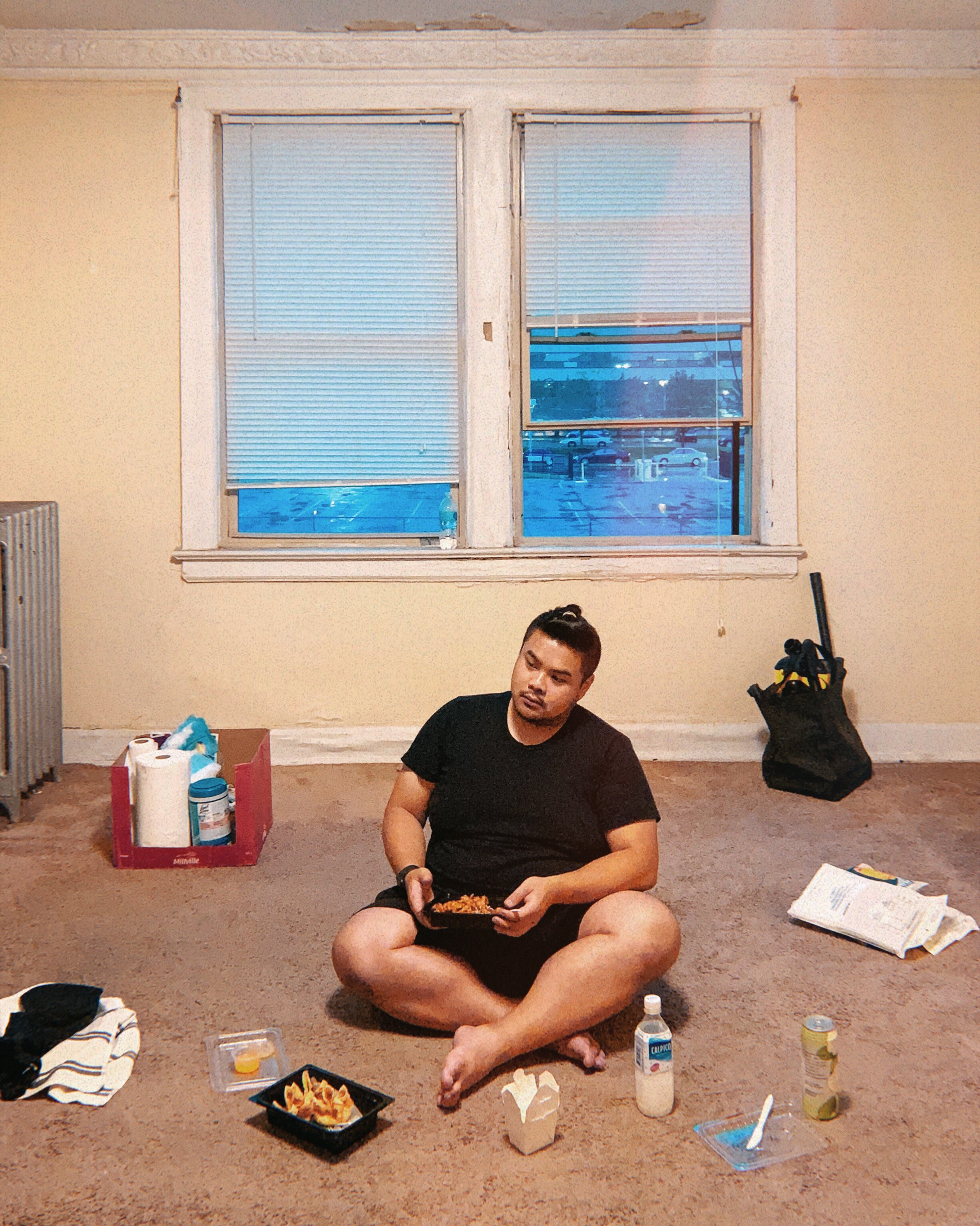RV Mendoza sits on the floor eating in a mostly empty apartment while moving supplies surround him.