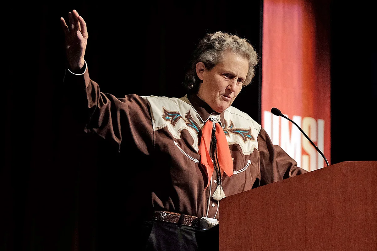 Temple Grandin raises one hand while speaking at a podium.