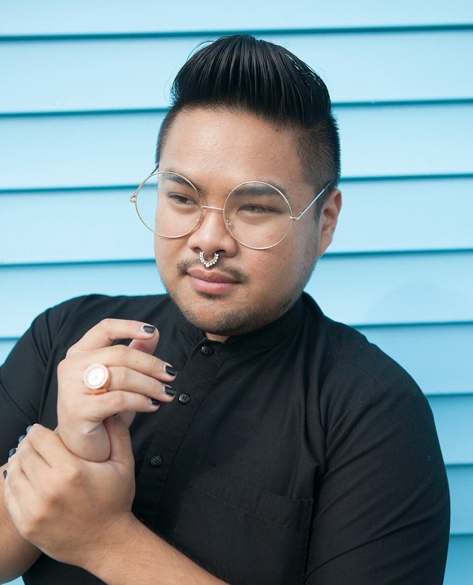 RV Mendoza poses in black clothing and round glasses against pale blue siding.